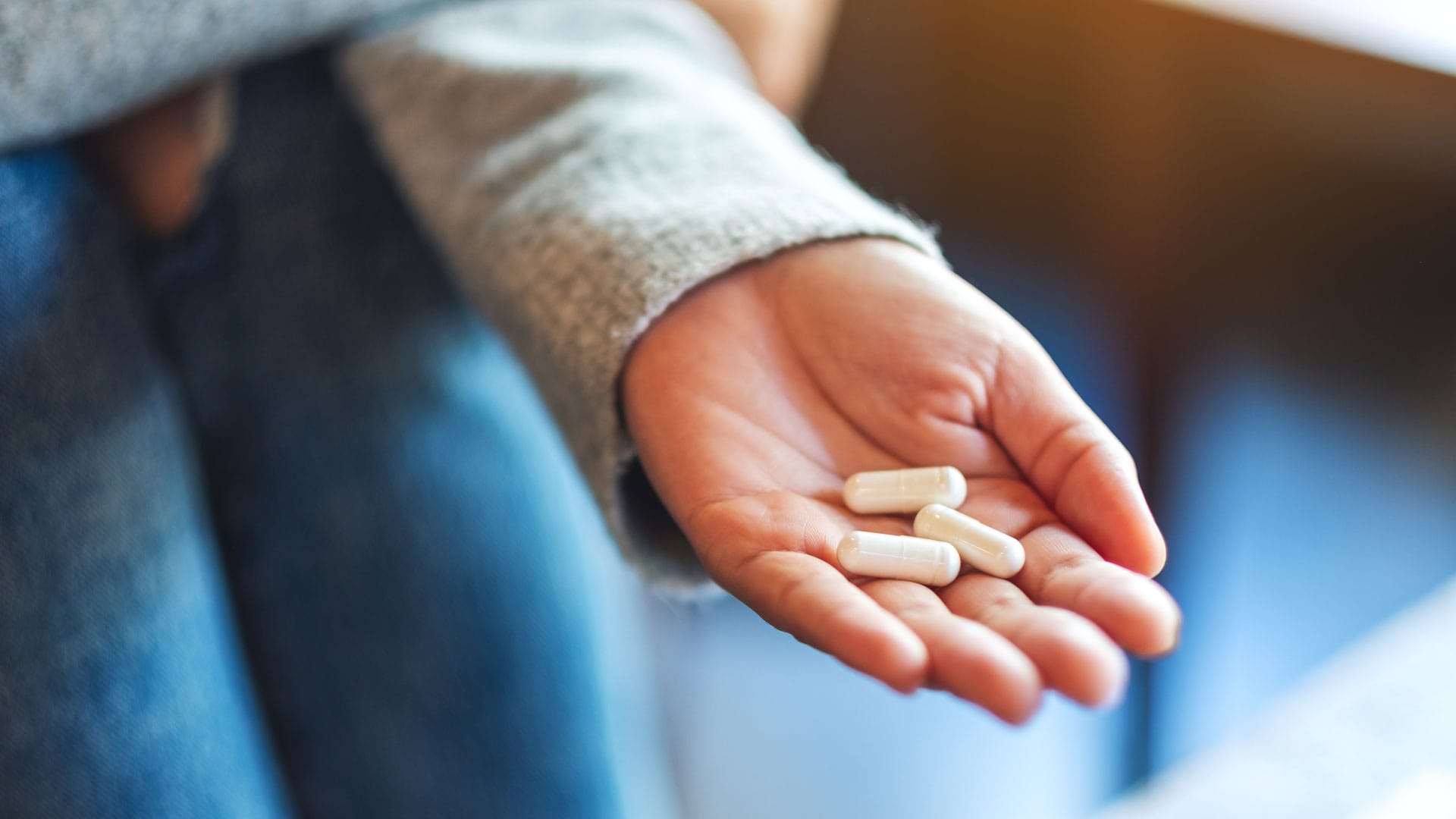 Bilden visar en person med blå trasiga jeans och ljusblå långärmad tröja som sträcker fram en hand med tre medicin kapslar i.